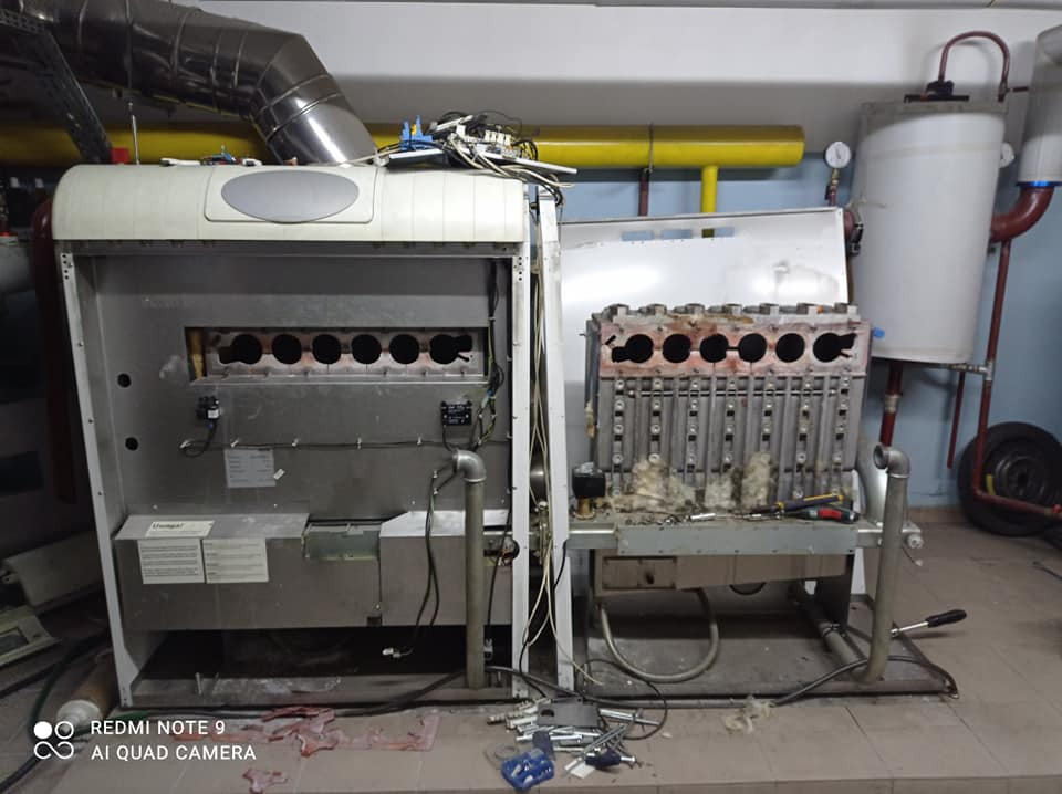 Rozłożony piec centralnego ogrzewania podczas serwisu, widoczne elementy grzewcze, narzędzia, instalacja hydrauliczna i fragment kotłowni.