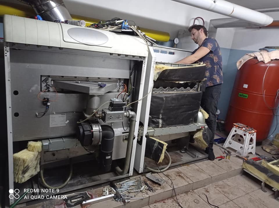 Mężczyzna w trakcie prac serwisowych przy otwartym piecu grzewczym, widoczne elementy izolacyjne, narzędzia na podłodze i czerwony zbiornik wyrównawczy.