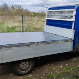 Niebieski bus z otwartą platformą ładunkową na tle łąki z drzewami, widoczne ślady użytkowania na karoserii.