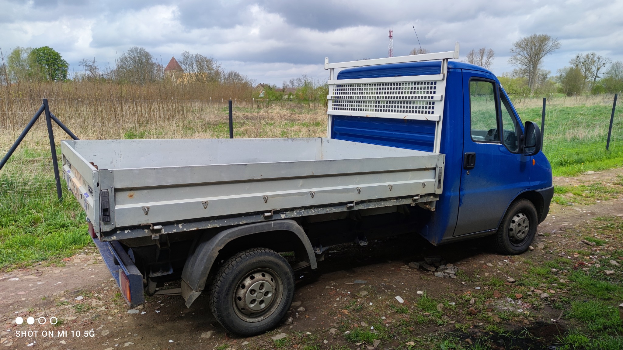 Niebieski samochód dostawczy z otwartą skrzynią ładunkową, zaparkowany na ziemi, z widocznym budynkiem w oddali pod pochmurnym niebem.