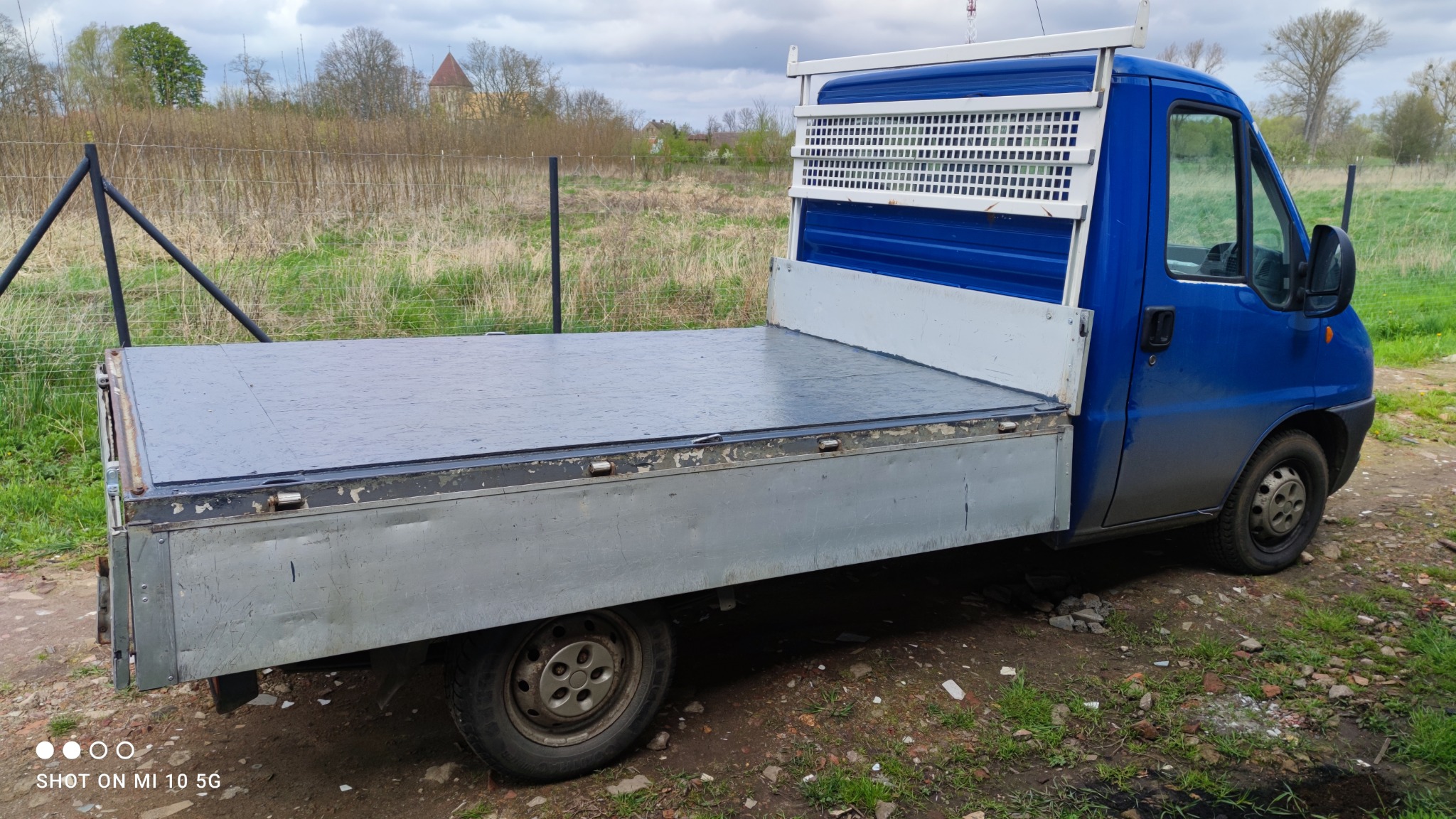 Niebieski bus z otwartą platformą ładunkową na tle łąki z drzewami, widoczne ślady użytkowania na karoserii.