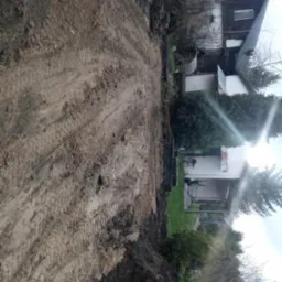 Wyrównany teren pod budowę domu jednorodzinnego, widoczne ślady opon ciężkiego sprzętu, w tle budynek mieszkalny i roślinność.