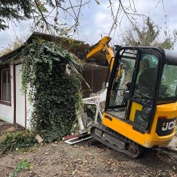 Usługi minikoparka - Żółta mini koparka JCB częściowo rozbiera biały budynek porośnięty bluszczem, widoczne uszkodzenia dachu i ściany, operator w kabinie.