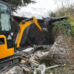 Żółta mini koparka podczas rozbiórki budynku, widoczne gruzy i zarośla