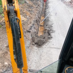 Usługi minikoparka - Wykopany wąski rów pod instalacje, widok z kabiny koparki, z boku widoczny fragment ramienia koparki, na dnie rowu pomarańczowy element instalacji, w tle zaparkowany samochód osobowy z otwartym...