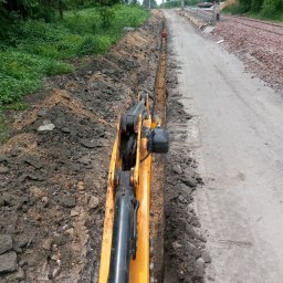 Usługi minikoparka - Żółta koparka tworzy wąski wykop wzdłuż drogi gruntowej, widoczne kable w wykopie, zarośla i linia kolejowa w tle.