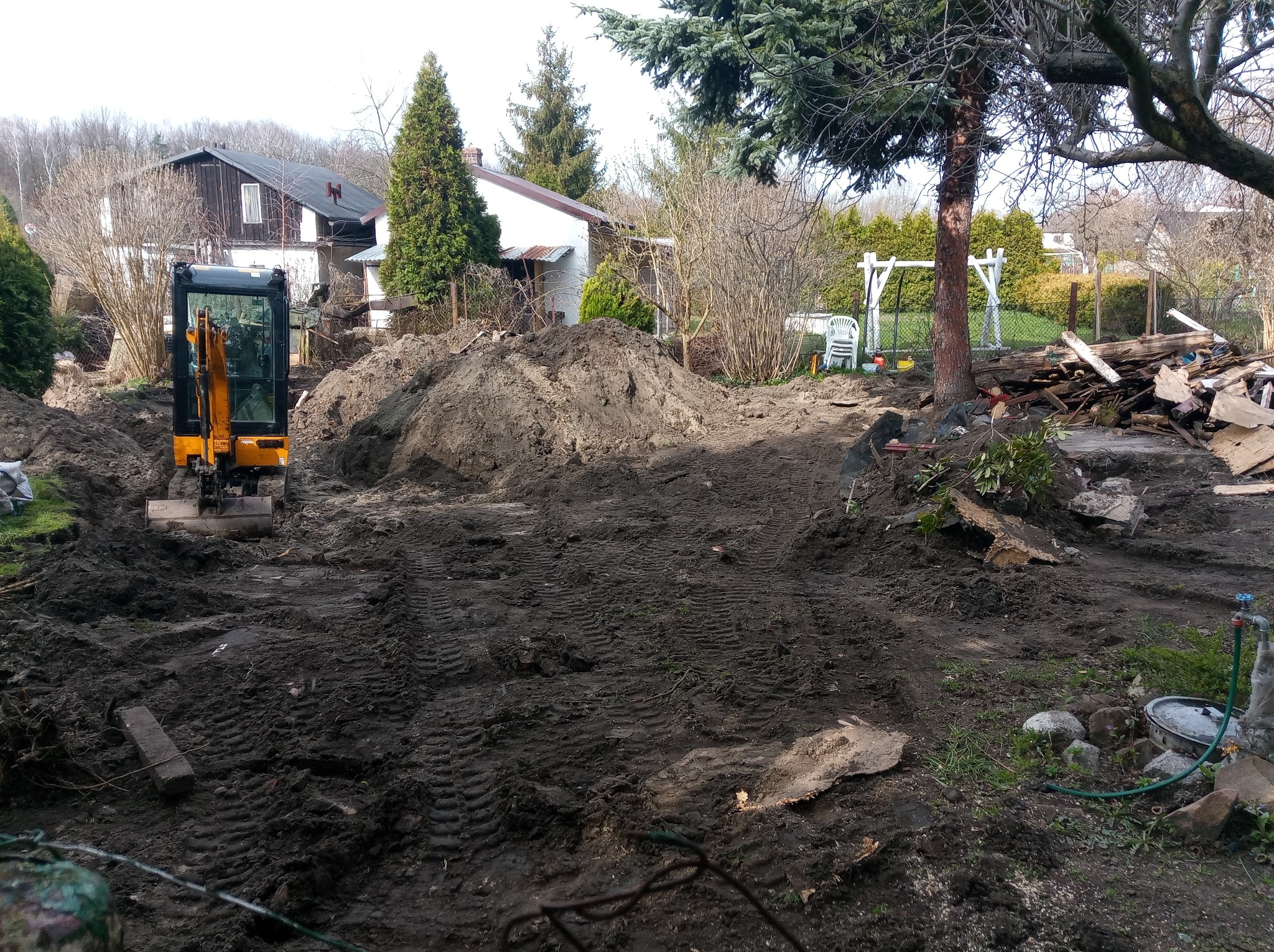 Mała koparka na błotnistym terenie budowy, w tle domy jednorodzinne i sterta gruzu, widoczne ślady opon na ziemi.