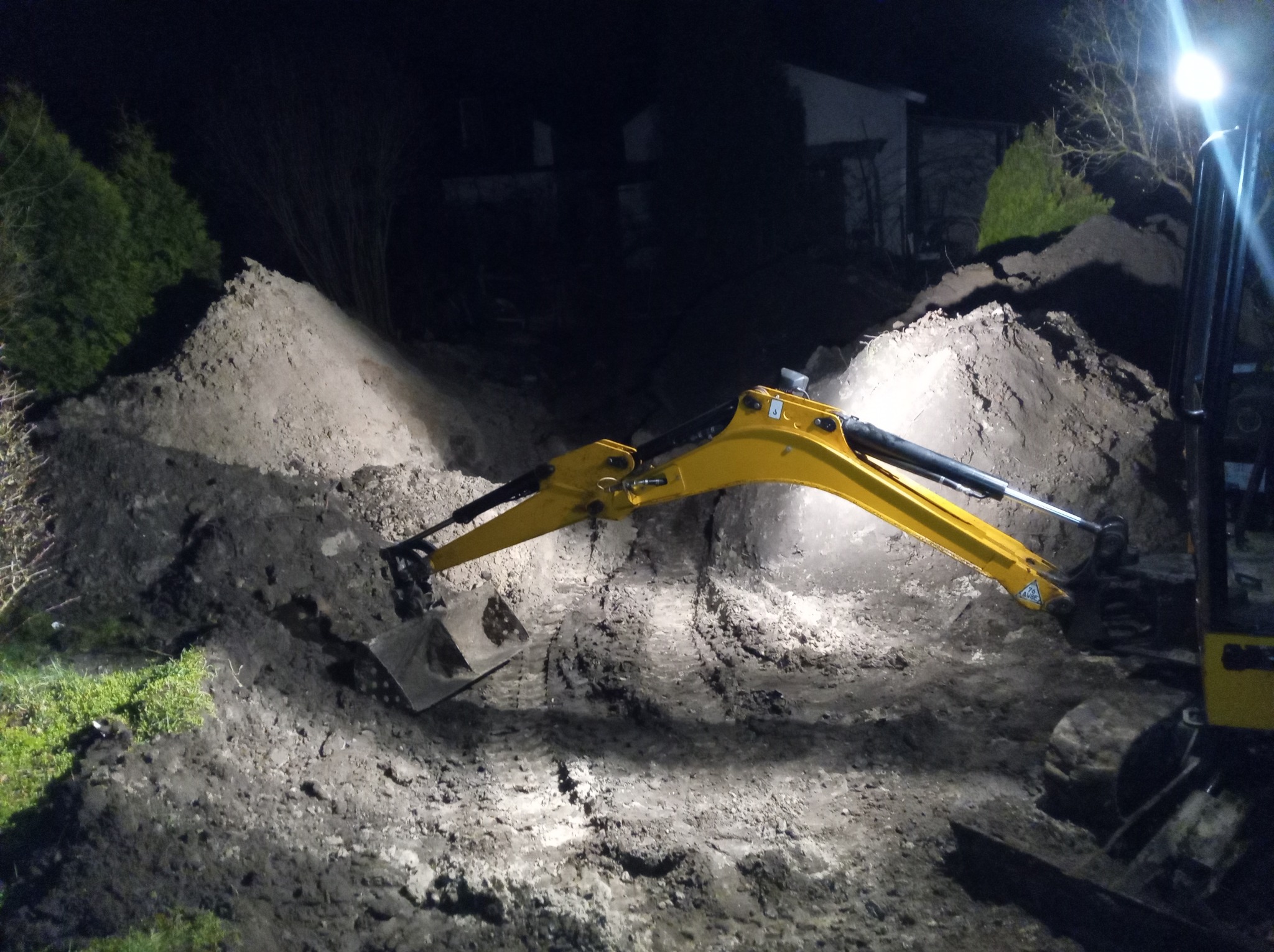 Żółta mini koparka z łyżką w wykopie, oświetlona reflektorem, pryzmy ziemi w tle, nocna sceneria.