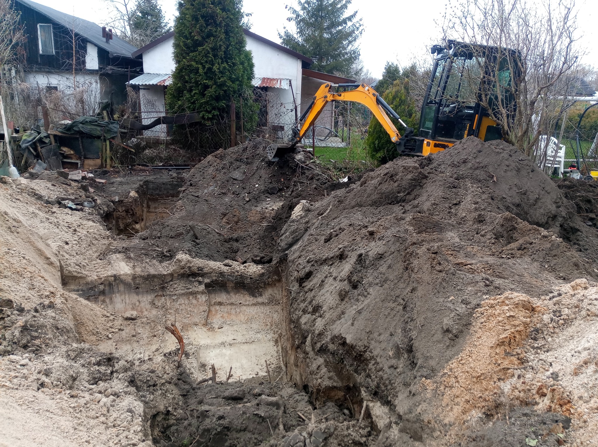 Żółta koparka wykonuje wykop w ziemi w Rudzie Śląskiej, widoczne ślady pracy i pryzmy ziemi, w tle zabudowania mieszkalne i drzewa.