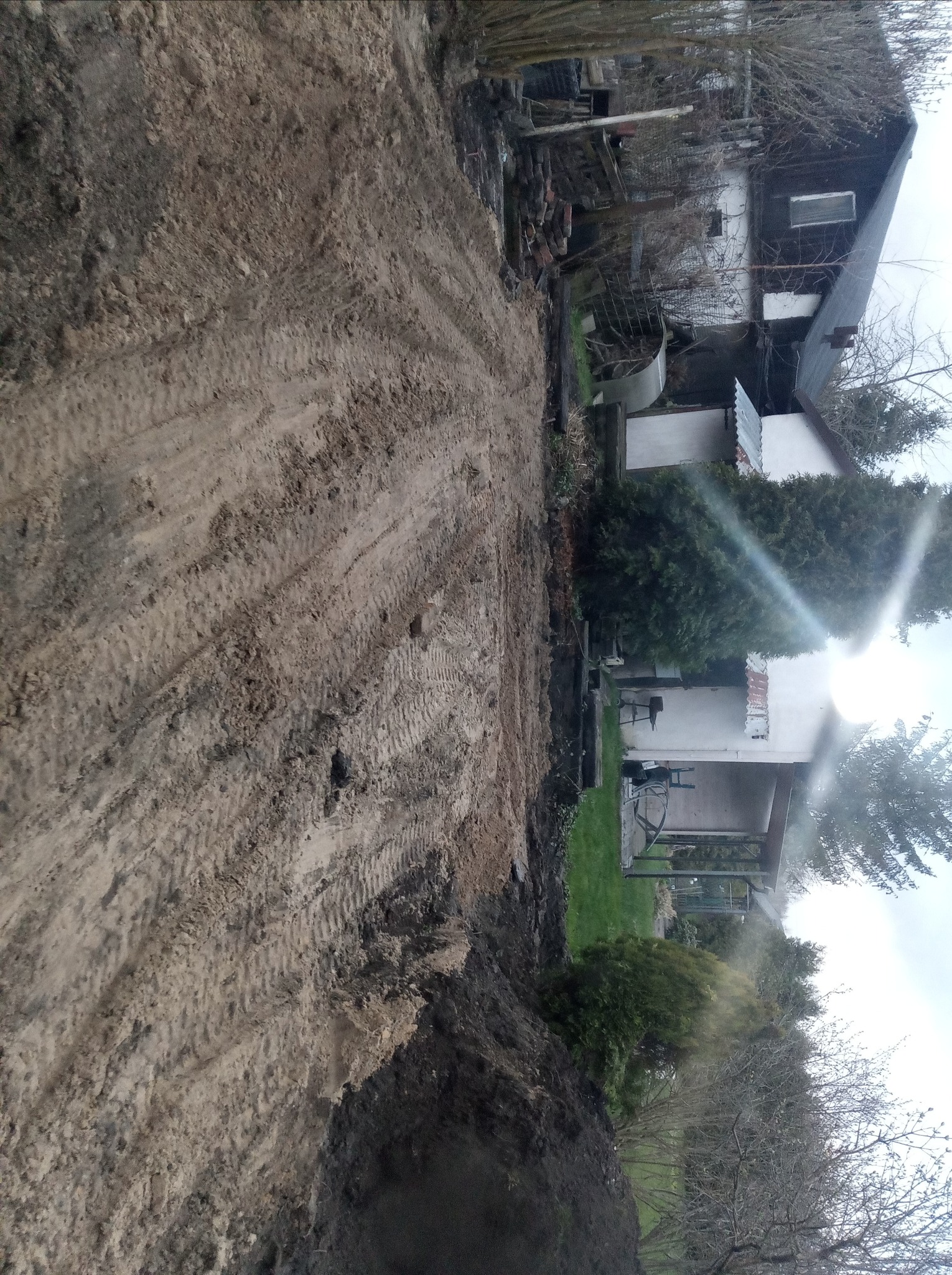 Wyrównany teren pod budowę domu jednorodzinnego, widoczne ślady opon ciężkiego sprzętu, w tle budynek mieszkalny i roślinność.