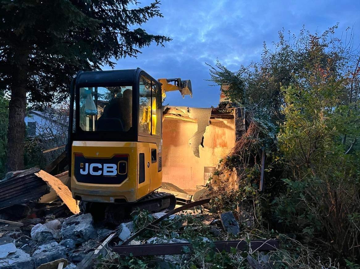 Żółta minikoparka JCB na gruzowisku podczas rozbiórki budynku, widoczne fragmenty muru i roślinność w tle, wieczorne oświetlenie.