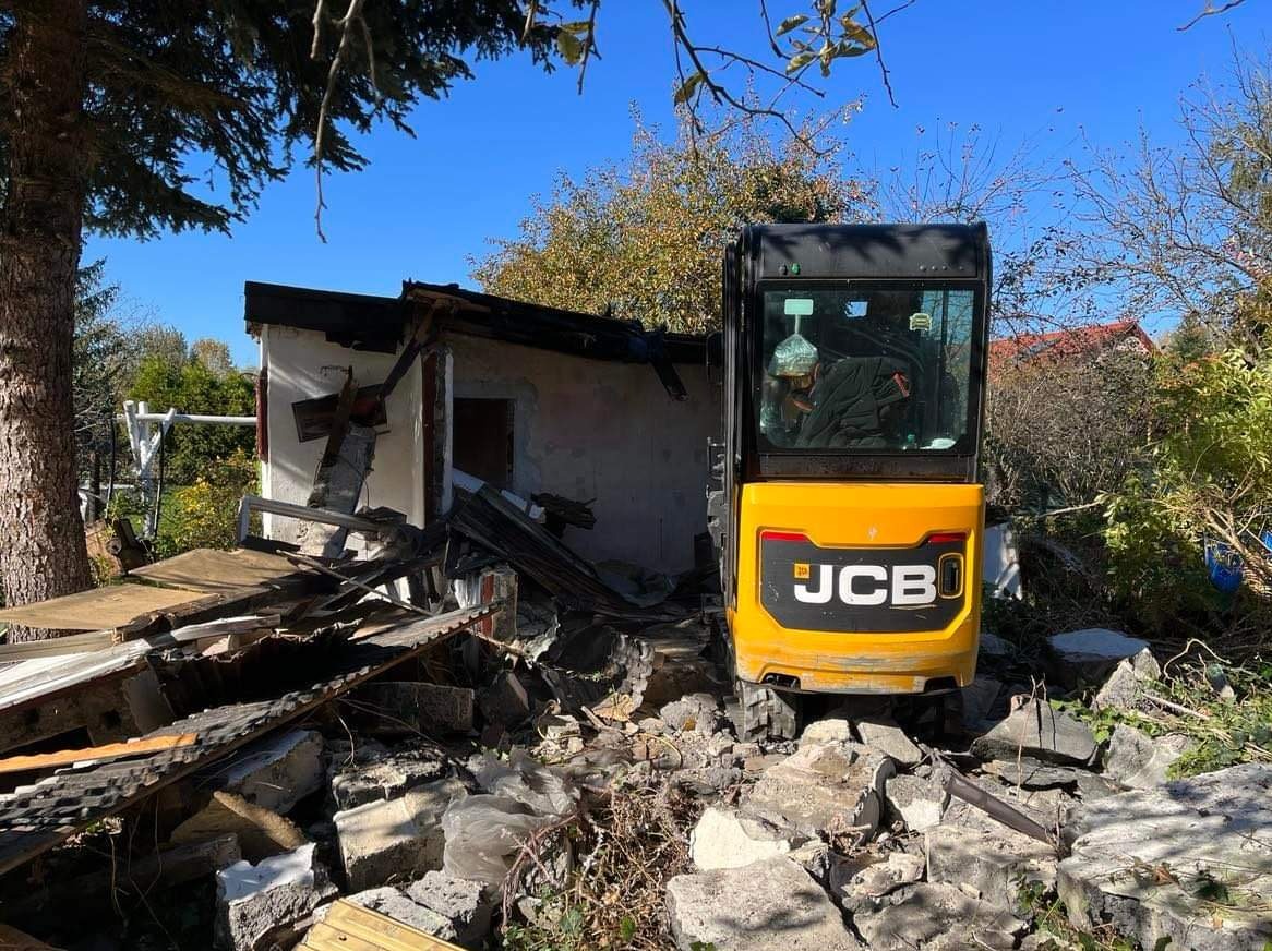 Żółta mini koparka JCB na tle częściowo zburzonego budynku z gruzem, drzewa i niebieskie niebo w tle, widok z tyłu maszyny.