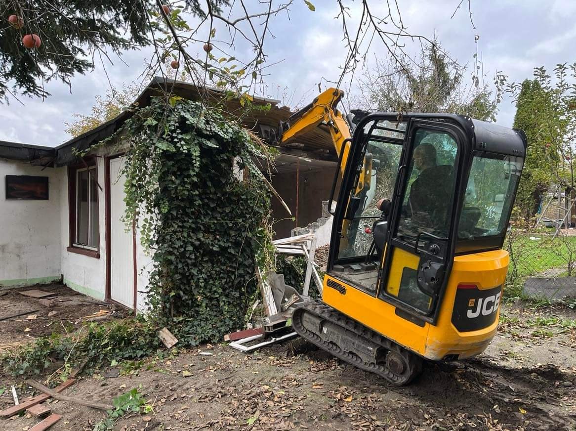 Żółta mini koparka JCB częściowo rozbiera biały budynek porośnięty bluszczem, widoczne uszkodzenia dachu i ściany, operator w kabinie.