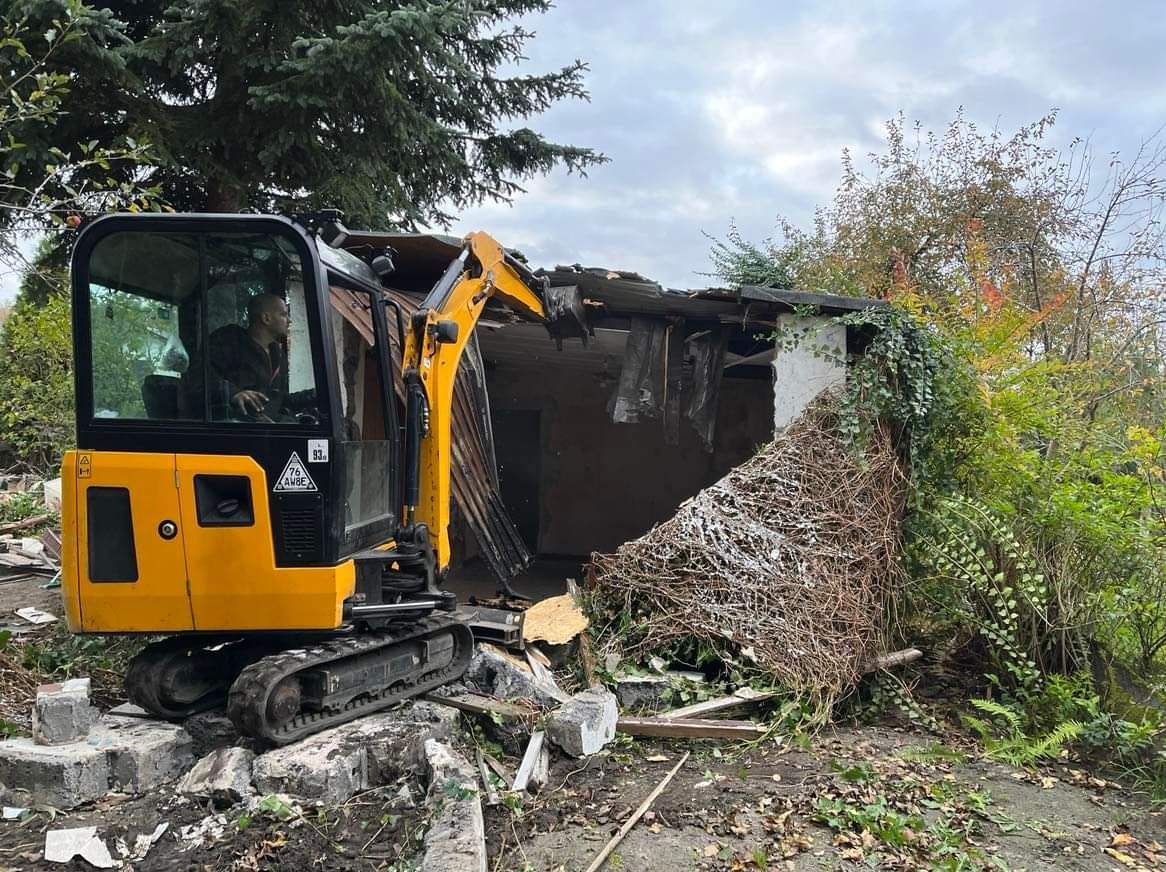 Żółta mini koparka gąsienicowa w trakcie pracy przy rozbiórce małego, zarośniętego budynku; operator w kabinie, gruz i zieleń wokół.