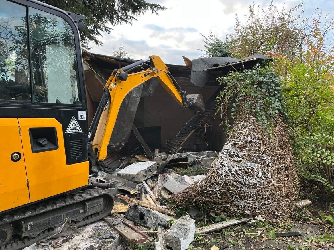 Żółta mini koparka podczas rozbiórki budynku, widoczne gruzy i zarośla