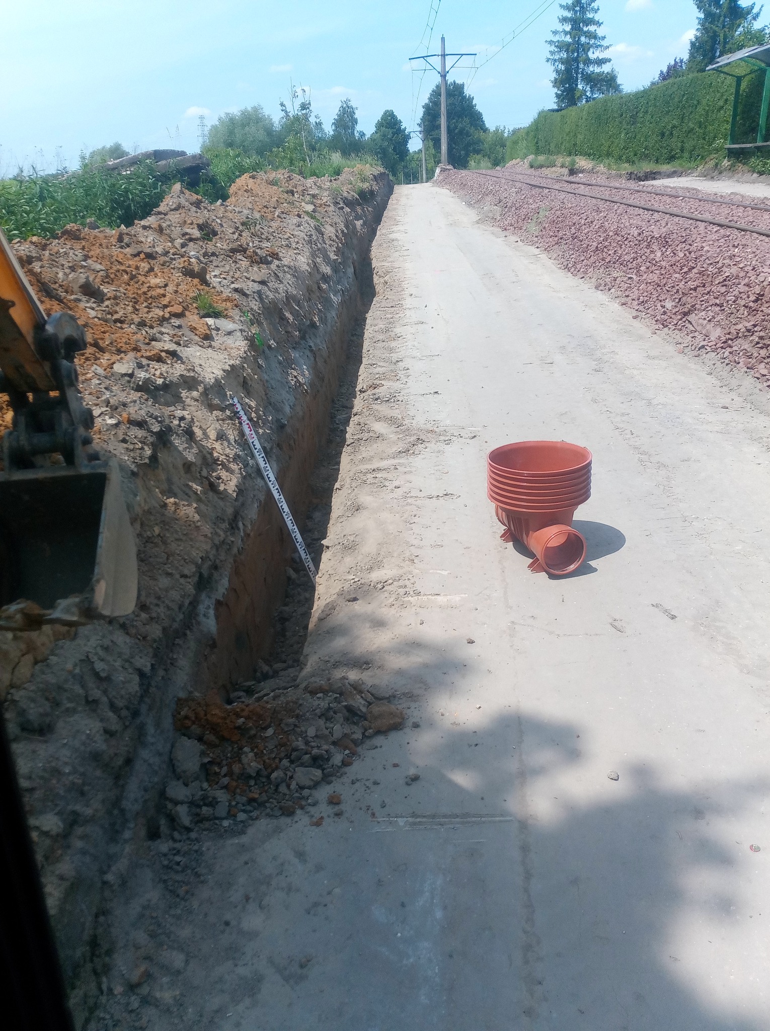Wąski wykop wzdłuż torów kolejowych, z widoczną łopatą koparki z lewej strony i ułożonymi rurami drenarskimi z prawej, obok leży miarka.