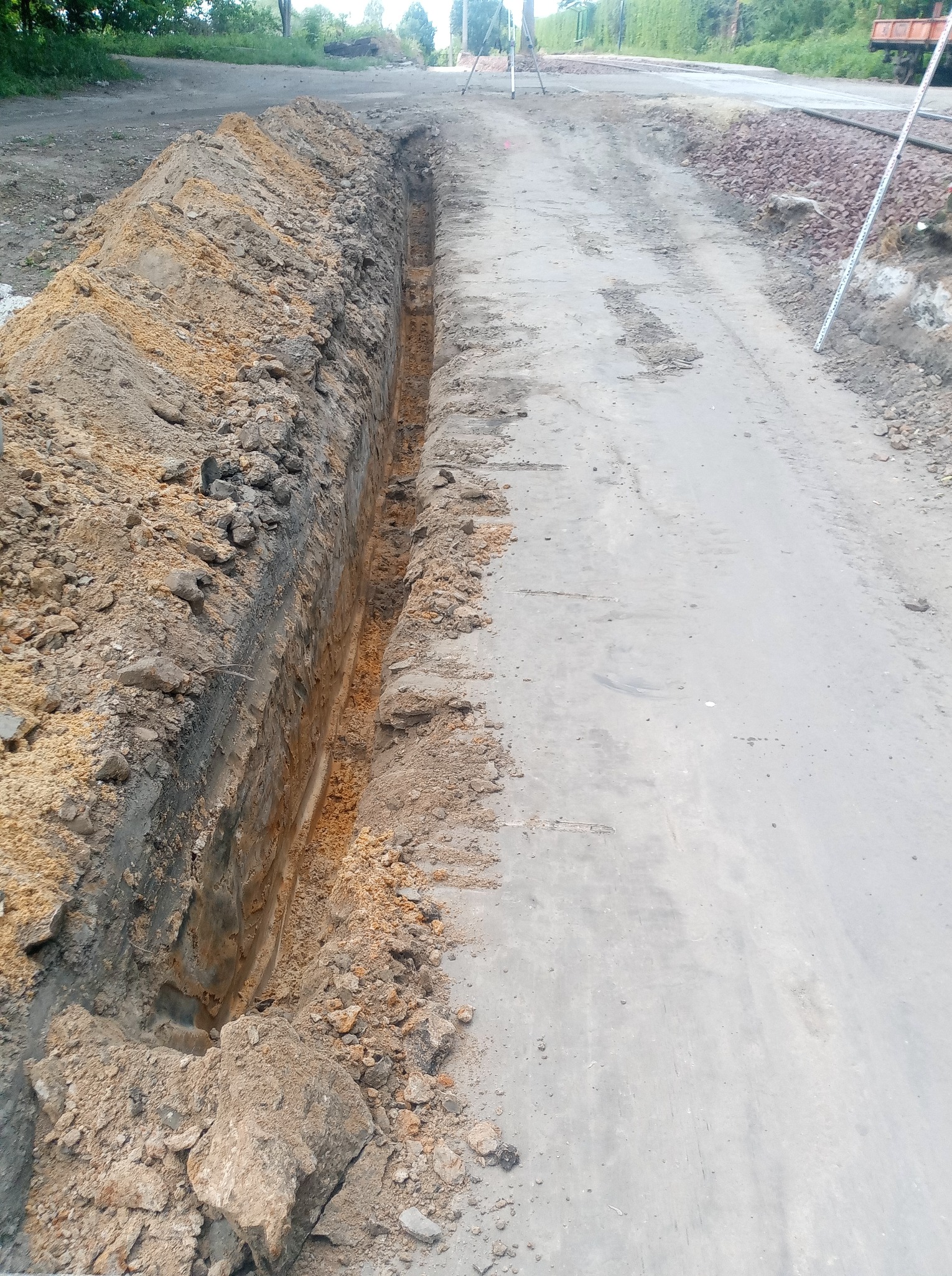 Wąski wykop w ziemi, prawdopodobnie pod instalacje, z hałdą ziemi po jednej stronie, na tle utwardzonej drogi w słoneczny dzień.
