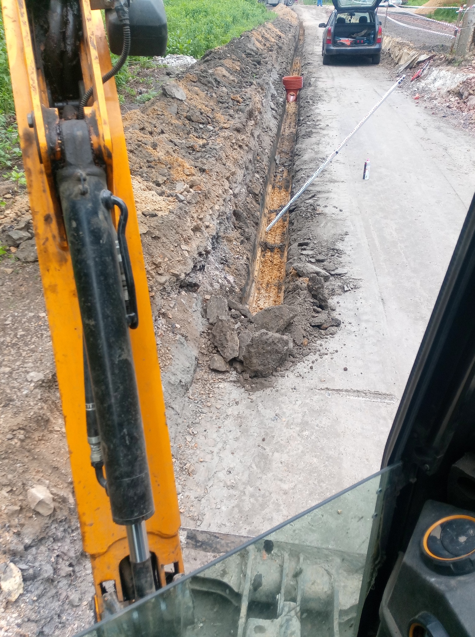 Wykopany wąski rów pod instalacje, widok z kabiny koparki, z boku widoczny fragment ramienia koparki, na dnie rowu pomarańczowy element instalacji, w tle zaparkowany samochód osobowy z otwartym...