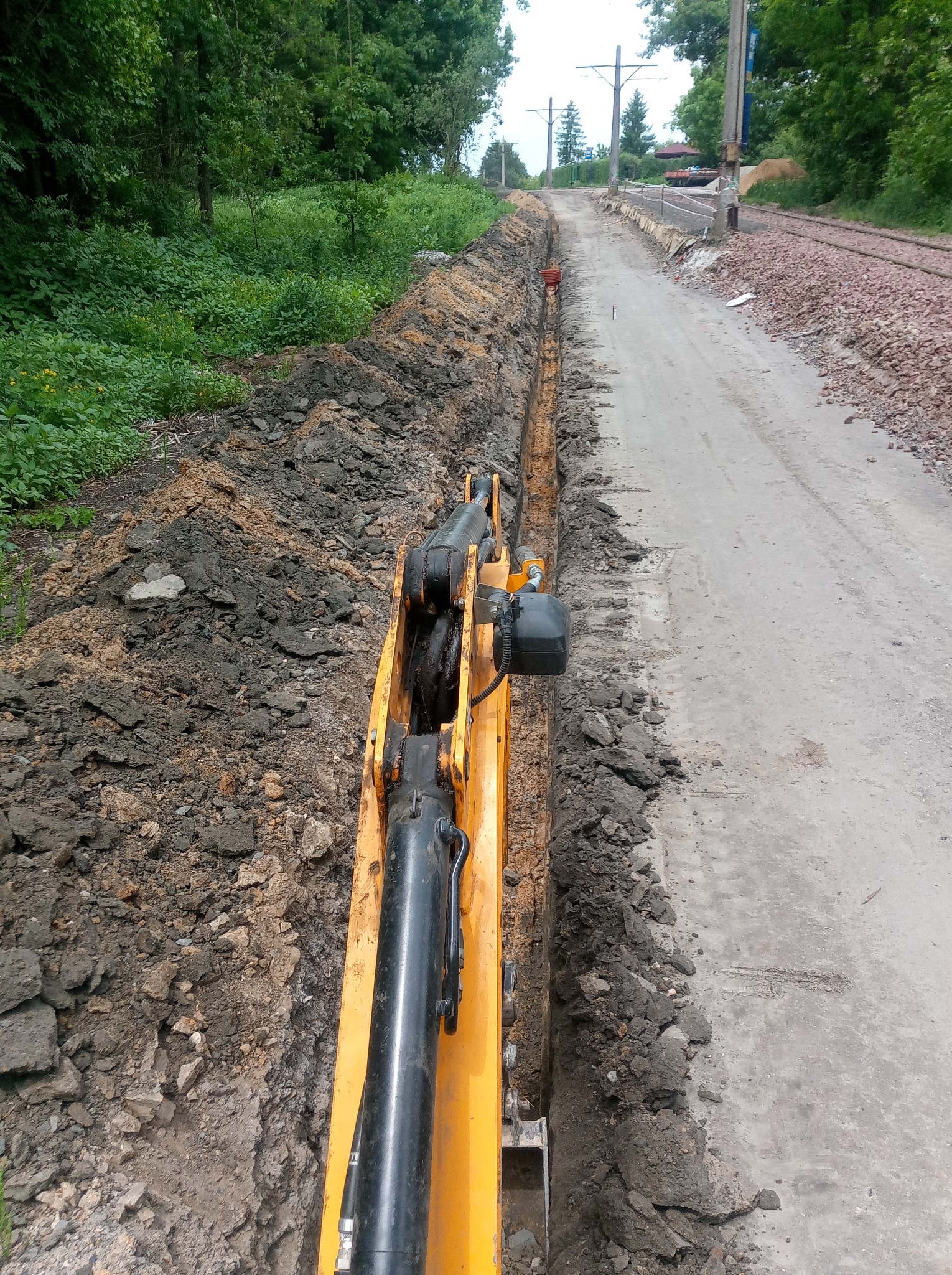 Żółta koparka tworzy wąski wykop wzdłuż drogi gruntowej, widoczne kable w wykopie, zarośla i linia kolejowa w tle.