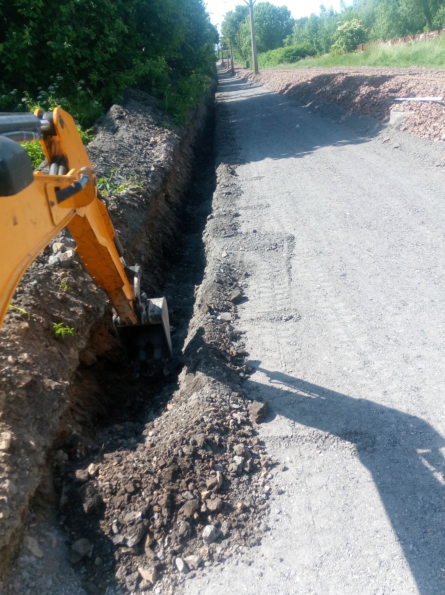 Żółta koparka wykonuje wykop wzdłuż drogi gruntowej, świeża ziemia usypana obok wykopu, w tle widoczna zieleń i drzewa.