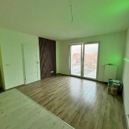 Fixit - Pusty pokój po remoncie z panelami podłogowymi o ciepłym odcieniu drewna, częściowo wykończony, z dekoracyjną ścianą z drewnianych listew, balkonem i pudełkami w rogu.