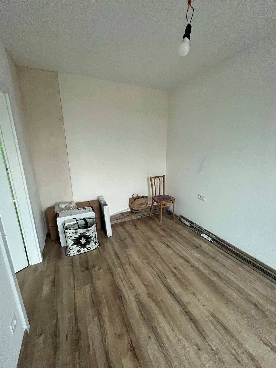 Pokój po remoncie z nową podłogą z paneli drewnopodobnych, widoczne kartony, grzejnik, krzesło i wisząca żarówka.
