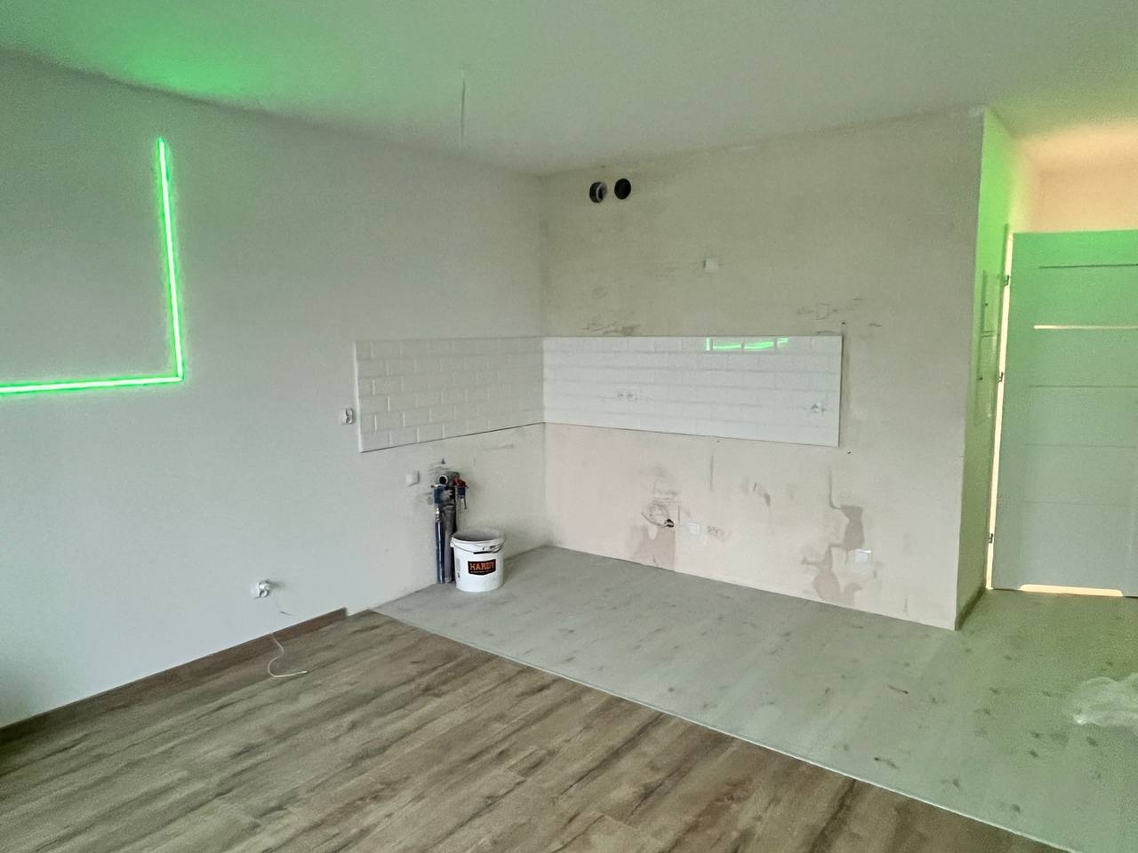 Fragment wnętrza w trakcie prac wykończeniowych: częściowo ułożona podłoga z paneli drewnopodobnych, fragment ściany z białymi płytkami typu metro, widoczne instalacje hydrauliczne i elektryczne...