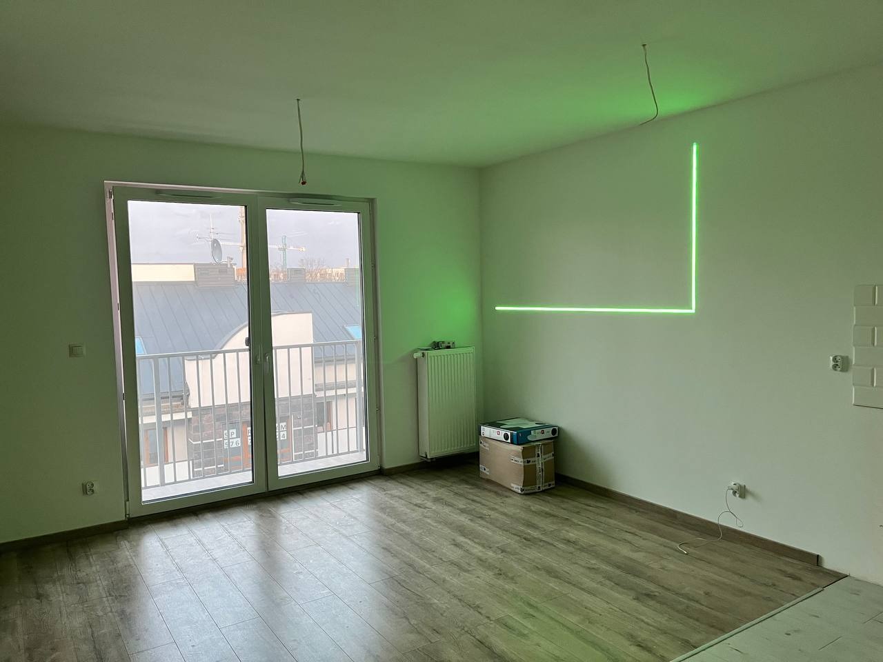 Pusty pokój z drewnopodobną podłogą, grzejnikiem i balkonem, poziomica laserowa wyświetla zielone linie na ścianie, przygotowanie do prac wykończeniowych.