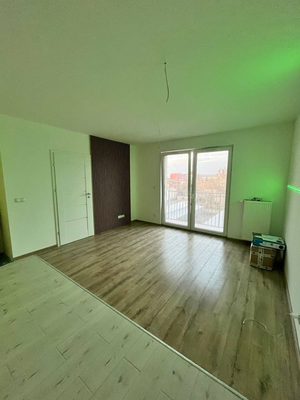 Pusty pokój po remoncie z panelami podłogowymi o ciepłym odcieniu drewna, częściowo wykończony, z dekoracyjną ścianą z drewnianych listew, balkonem i pudełkami w rogu.