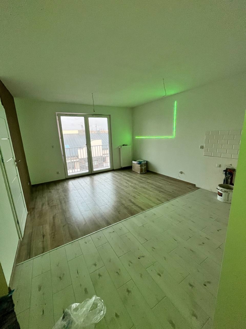 Wnętrze w trakcie prac wykończeniowych: częściowo ułożone panele podłogowe, na ścianie zielona linia lasera wyznaczająca poziom, widoczne wiadro z farbą i kartonowe pudło.