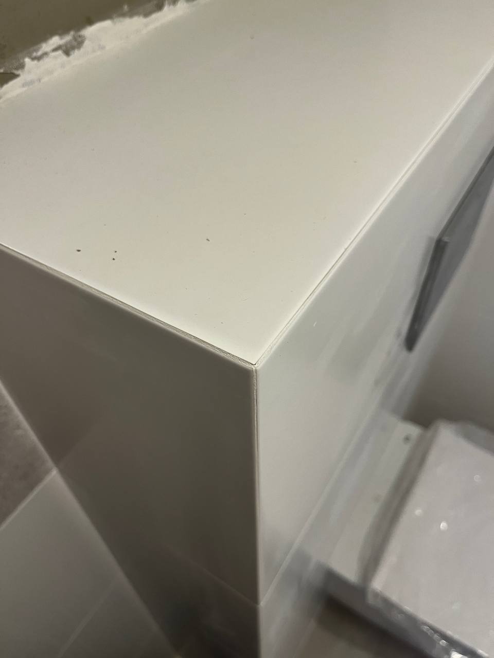 Białe, błyszczące płytki ceramiczne ułożone na narożniku, z widocznym minimalnym spoinowaniem i fragmentem szarej płyty w tle. Ujęcie z bliska, podkreślające precyzję wykonania.