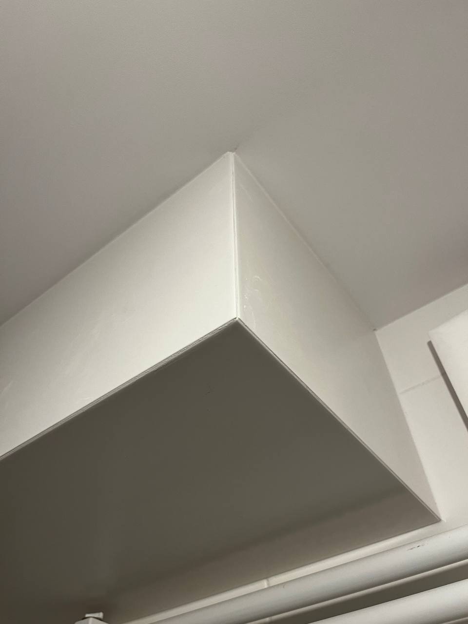 Nisko kątowe ujęcie pomalowanego na biało narożnika sufitu z widocznymi śladami świeżej farby i niedoskonałościami wykończenia, fragment rury grzewczej w tle.
