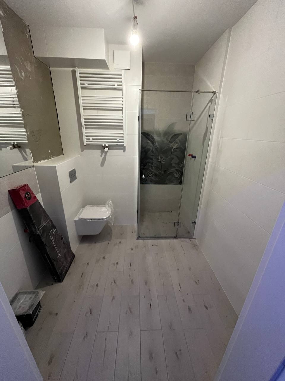 Nowoczesna łazienka w trakcie remontu, widoczna kabina prysznicowa ze wzorem roślinnym na tylnej ścianie, biała wisząca toaleta w folii ochronnej, grzejnik drabinkowy i podłoga wyłożona panelami...