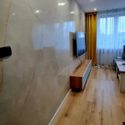 Ściana z efektem marmuru w salonie z drewnianą podłogą, wisząca szafka RTV i telewizor. Widoczne żółte zasłony i okno z białymi firankami w tle.