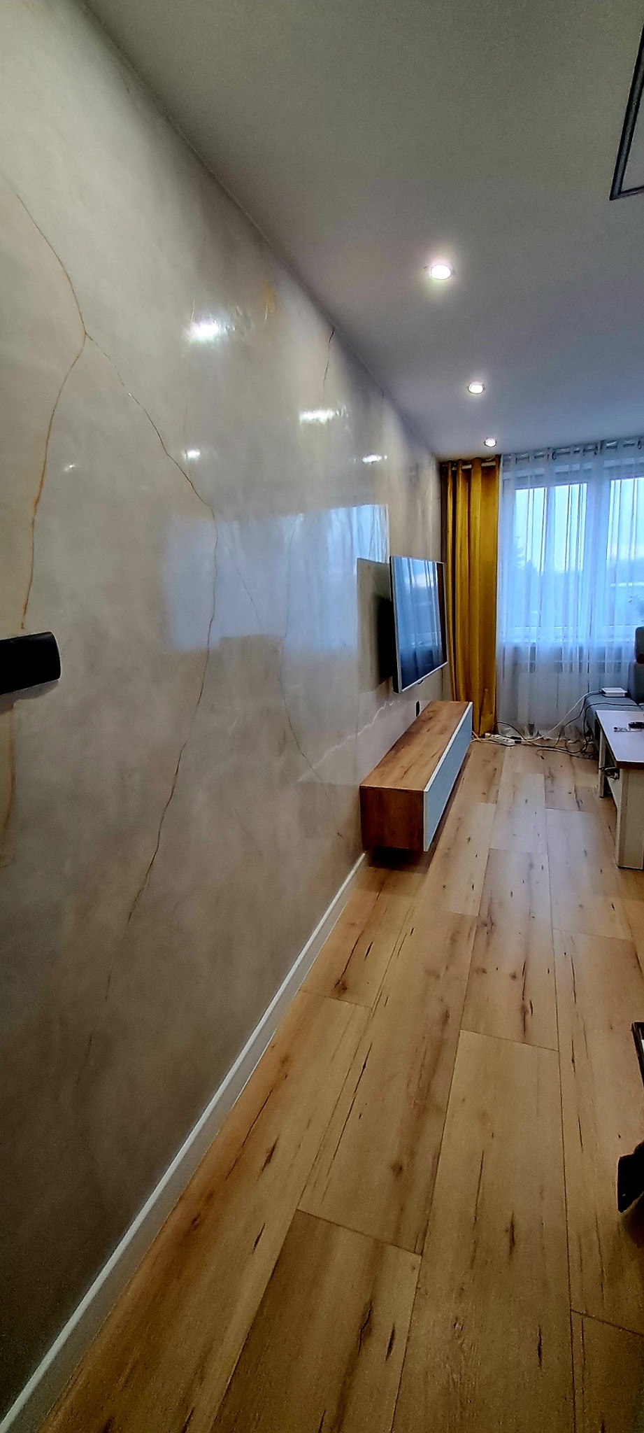 Ściana z efektem marmuru w salonie z drewnianą podłogą, wisząca szafka RTV i telewizor. Widoczne żółte zasłony i okno z białymi firankami w tle.