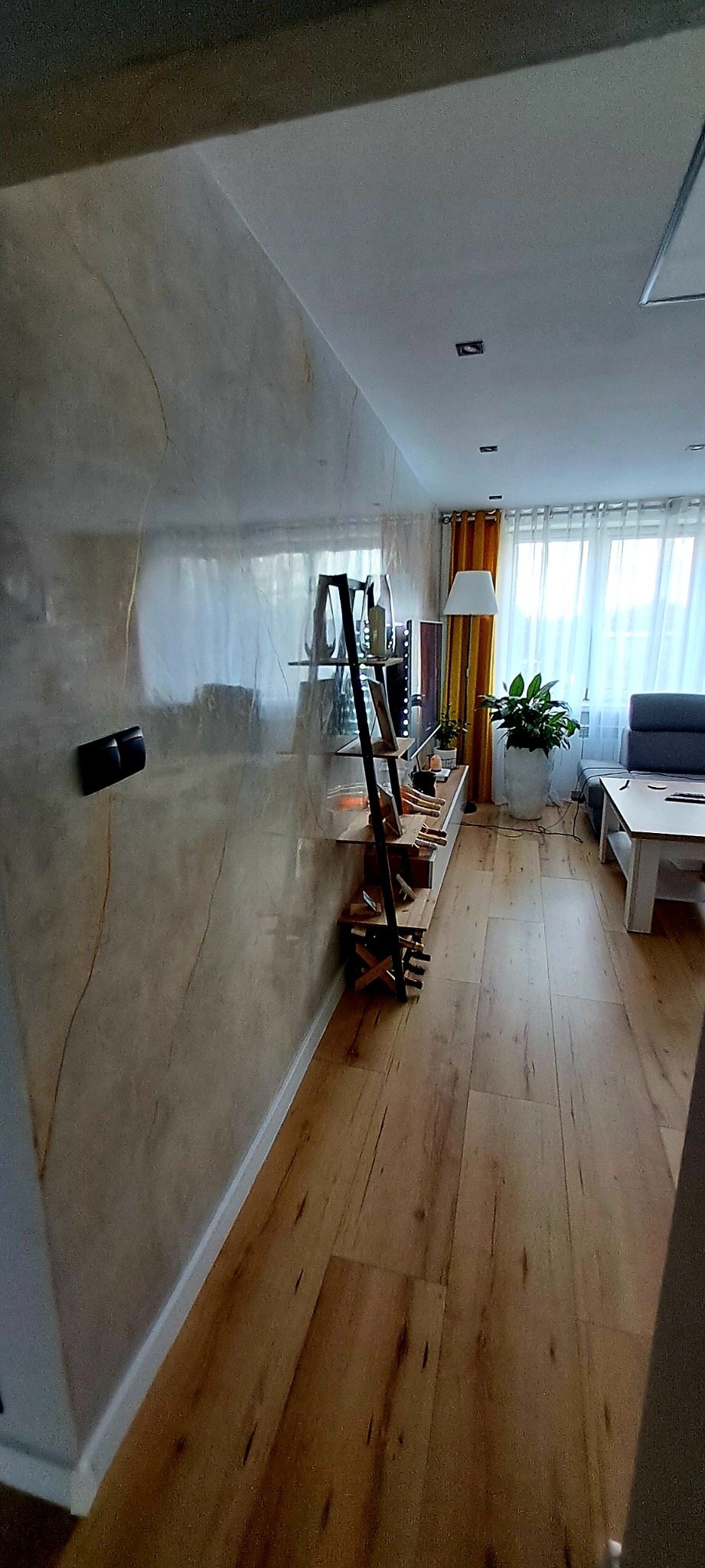 Ściana w salonie z efektem marmuru, drewniana podłoga, regał z elementami dekoracyjnymi i butelkami wina, widok na okno z zasłonami i fragment sofy.