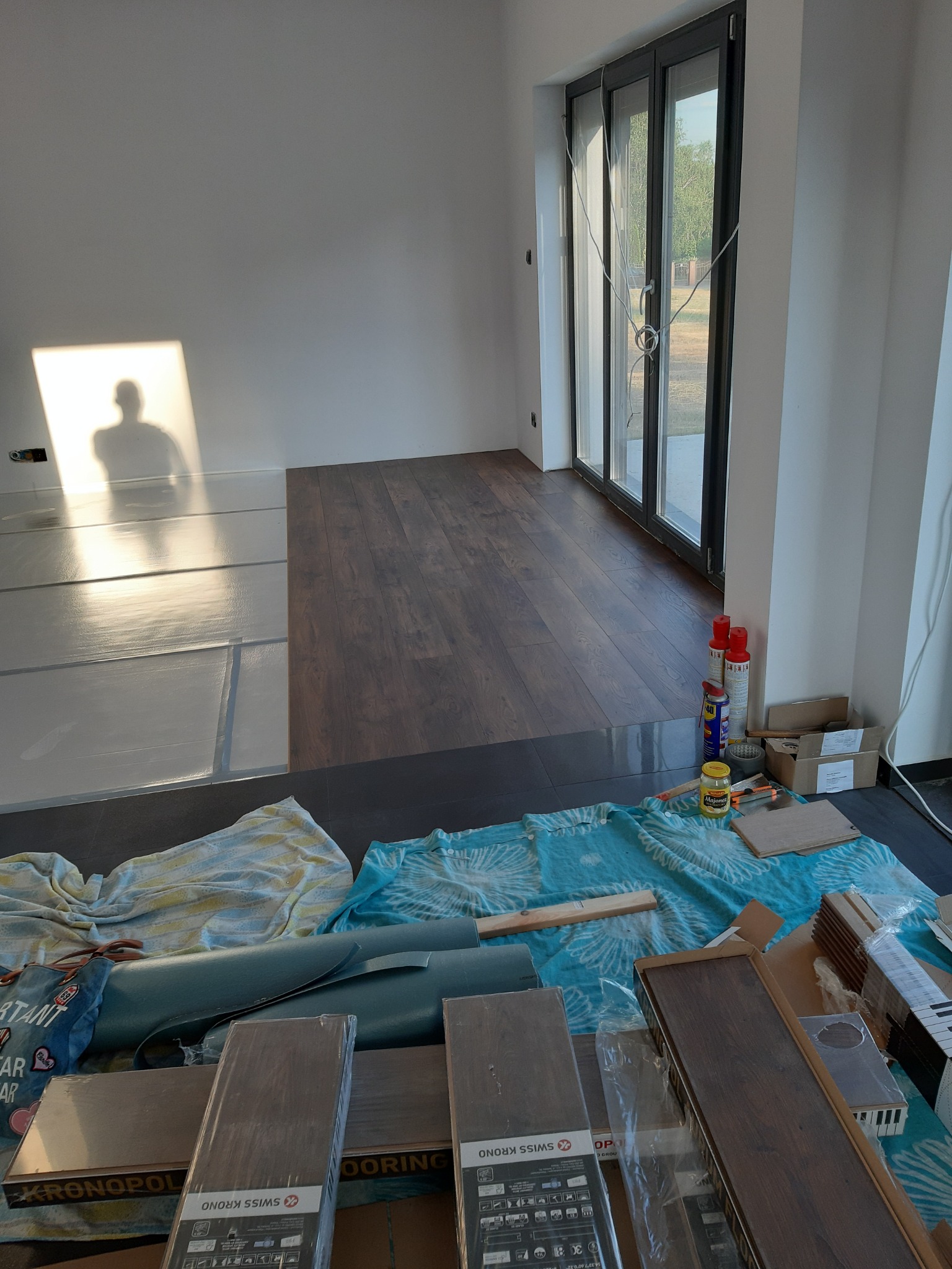 Proces układania paneli podłogowych w jasnym pomieszczeniu z dużym oknem tarasowym, widoczne paczki paneli Swiss Krono, podkład, narzędzia i cień osoby na ścianie.