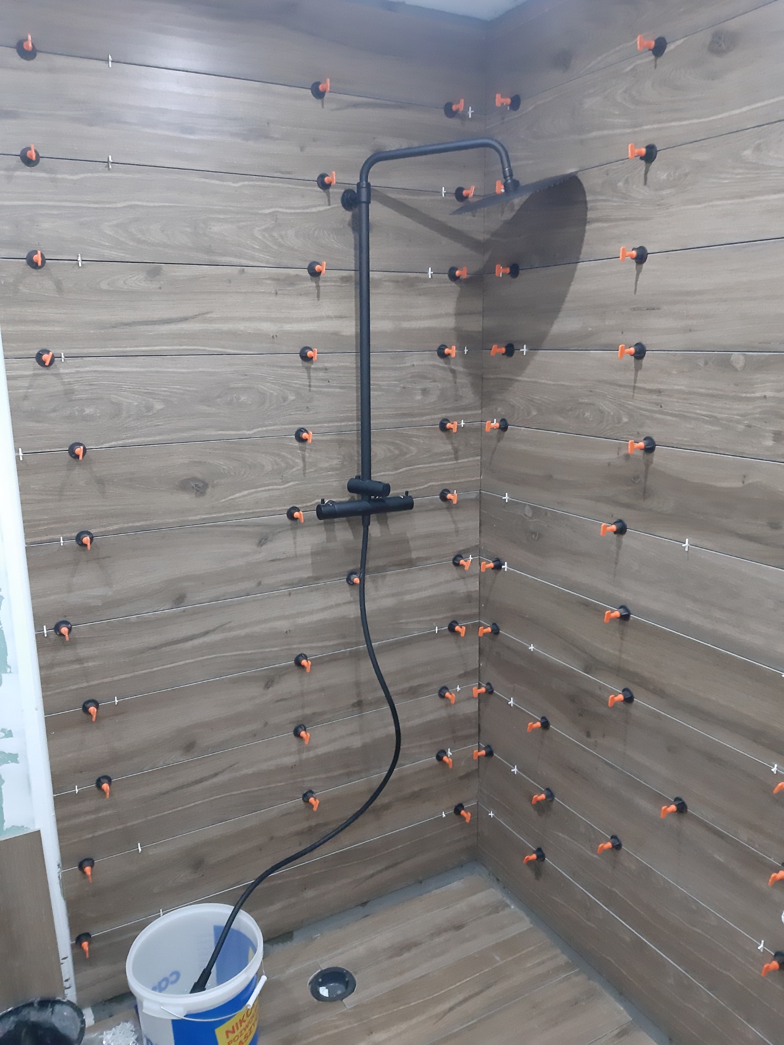 Proces układania płytek imitujących drewno w kabinie prysznicowej, z widocznymi klipsami poziomującymi i krzyżykami dystansowymi, oraz czarną armaturą prysznicową i wiaderkiem z resztkami kleju...