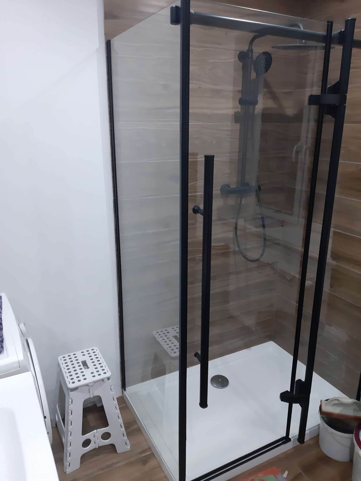 Nowoczesna kabina prysznicowa z czarnymi profilami i deszczownicą, zamontowana w łazience z drewnopodobnymi płytkami, widoczny biały brodzik i składany stołek.