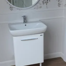 Nowoczesna łazienka z białą szafką pod umywalkę marki Kolo, okrągłe lustro i tapeta w kwiatowy wzór.