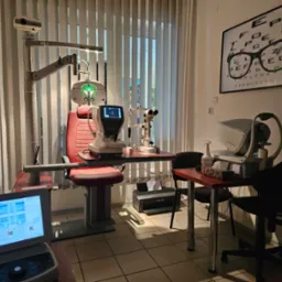 Gabinet lekarski z wyposażeniem: fotel pacjenta, lampa diagnostyczna, autorefraktometr, tonometr, tablica Snellena z okularami oraz monitor z wynikami pomiarów.