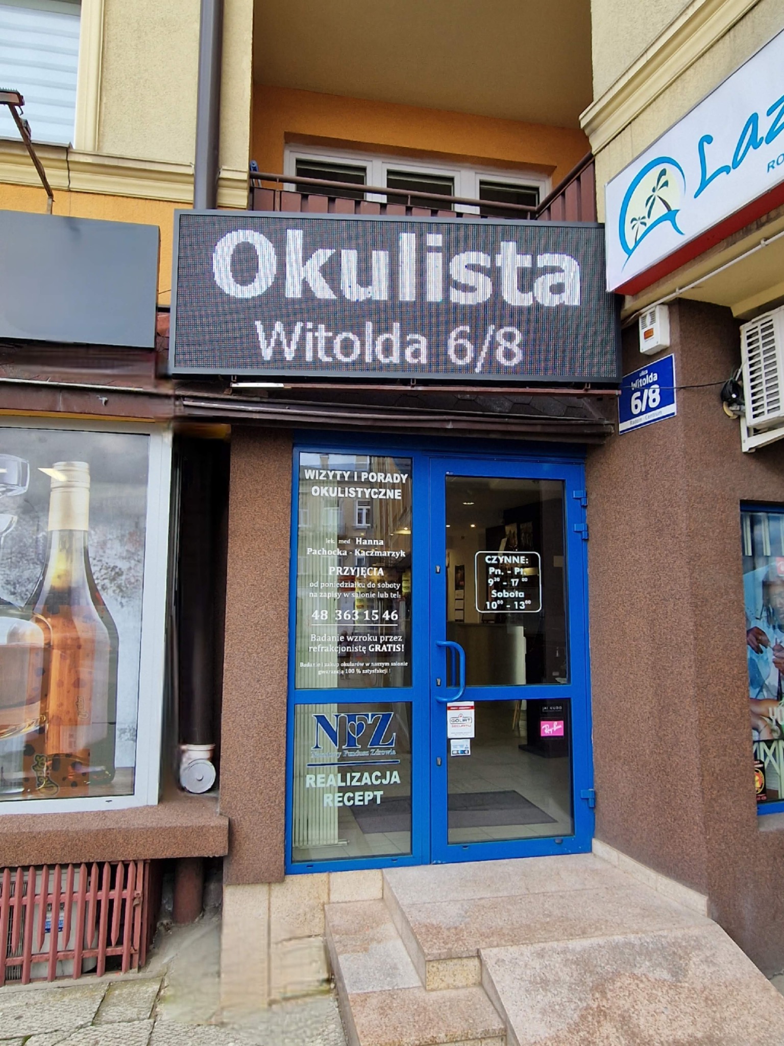 Fasada budynku z niebieskimi drzwiami wejściowymi do gabinetu z szyldem 'Okulista Witolda 6/8' nad wejściem, obok szyld z godzinami otwarcia i informacjami o badaniach wzroku.