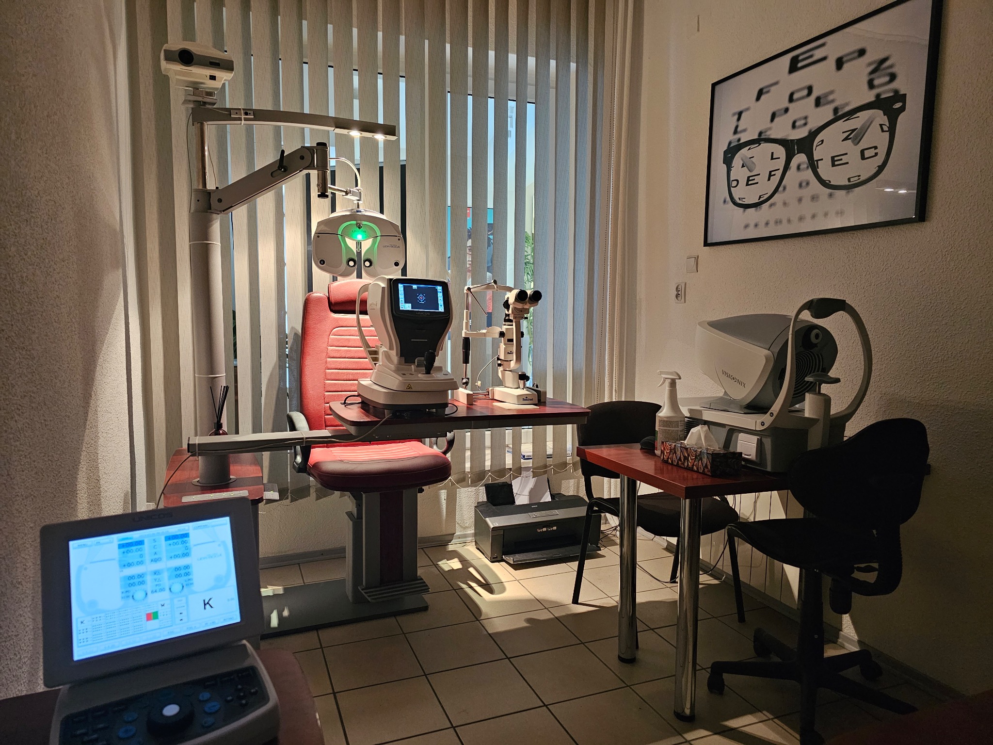 Gabinet lekarski z wyposażeniem: fotel pacjenta, lampa diagnostyczna, autorefraktometr, tonometr, tablica Snellena z okularami oraz monitor z wynikami pomiarów.