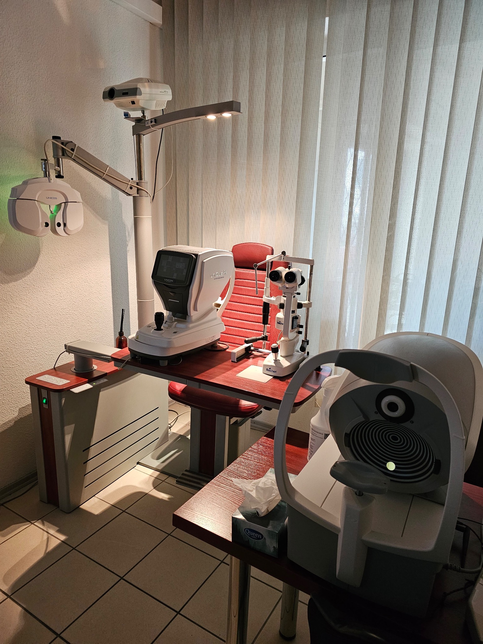 Wyposażenie gabinetu okulistycznego: foropter, autorefraktometr, biomikroskop i perymetr na tle pionowych żaluzji.