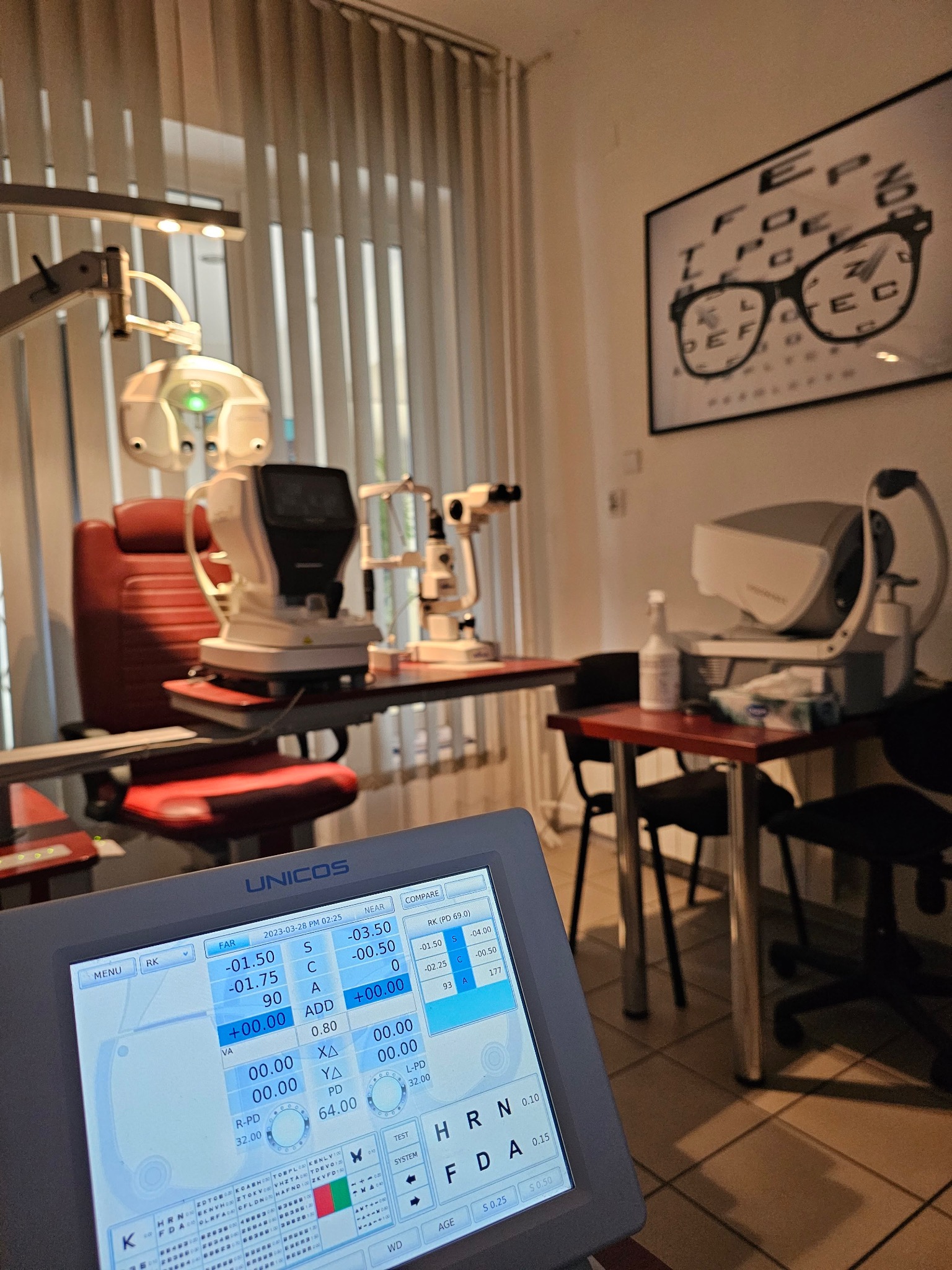 Gabinet okulistyczny z nowoczesnym sprzętem diagnostycznym, w tle tablica Snellena z okularami, na pierwszym planie ekran urządzenia z wynikami pomiarów.