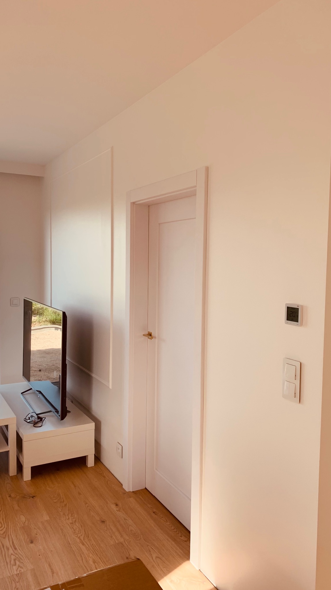 Jasne wnętrze po remoncie: drewniana podłoga, białe ściany z dekoracyjnymi listwami, białe drzwi z złotą klamką, telewizor na białej szafce, termostat na ścianie.