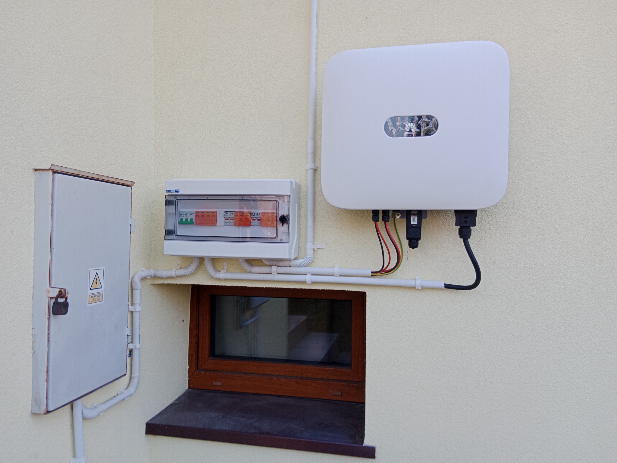 Zewnętrzna instalacja elektryczna: biała skrzynka rozdzielcza z widocznymi bezpiecznikami, metalowa skrzynka z symbolem ostrzegawczym i biały inwerter fotowoltaiczny zamontowany na elewacji budynku...