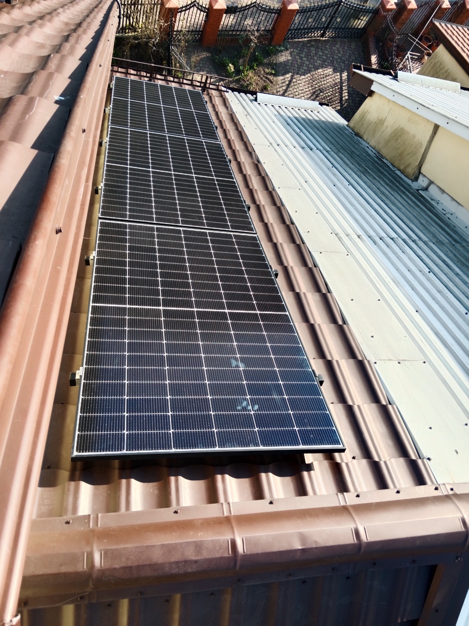 Dwa panele fotowoltaiczne zamontowane na brązowym, falistym dachu domu, widoczne z góry pod kątem, z fragmentem metalowego ogrodzenia w tle.