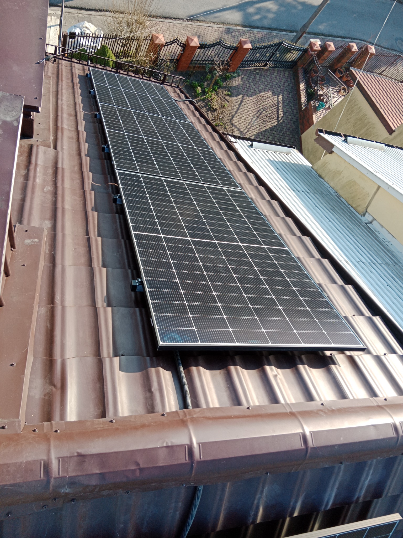 Widok z góry na panele fotowoltaiczne zamontowane na brązowym, metalowym dachu domu. Na tle widoczny fragment ulicy i sąsiednie budynki.