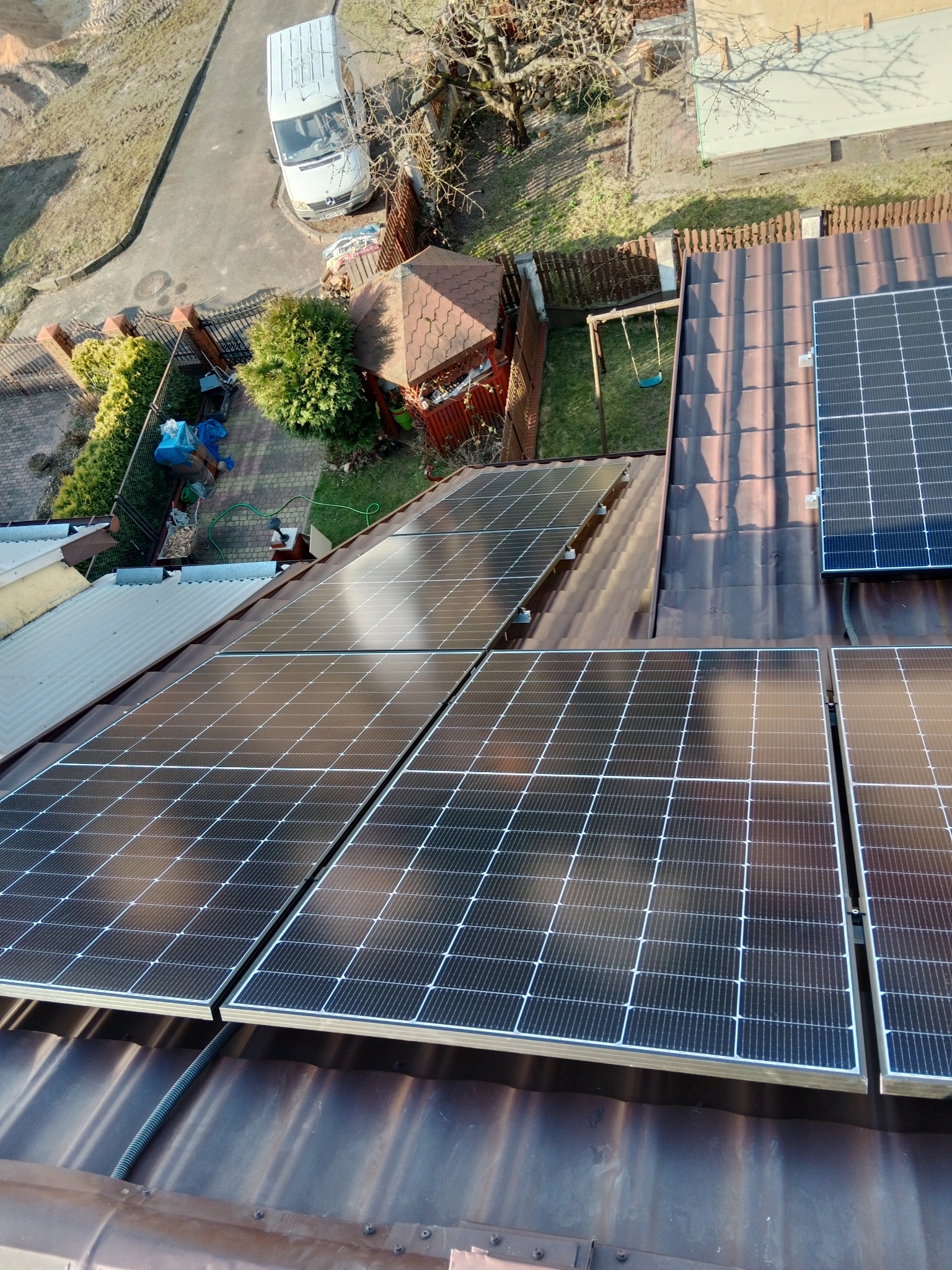 Widok z góry na brązowy dach pokryty panelami fotowoltaicznymi, na tle ogród z huśtawką, altaną i zaparkowany biały bus.